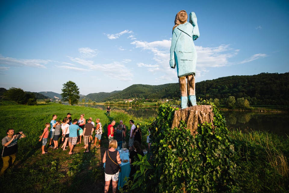 A light blue wooden statue of Melania Trump stands atop a tree stump in a field.  People gather nearby.