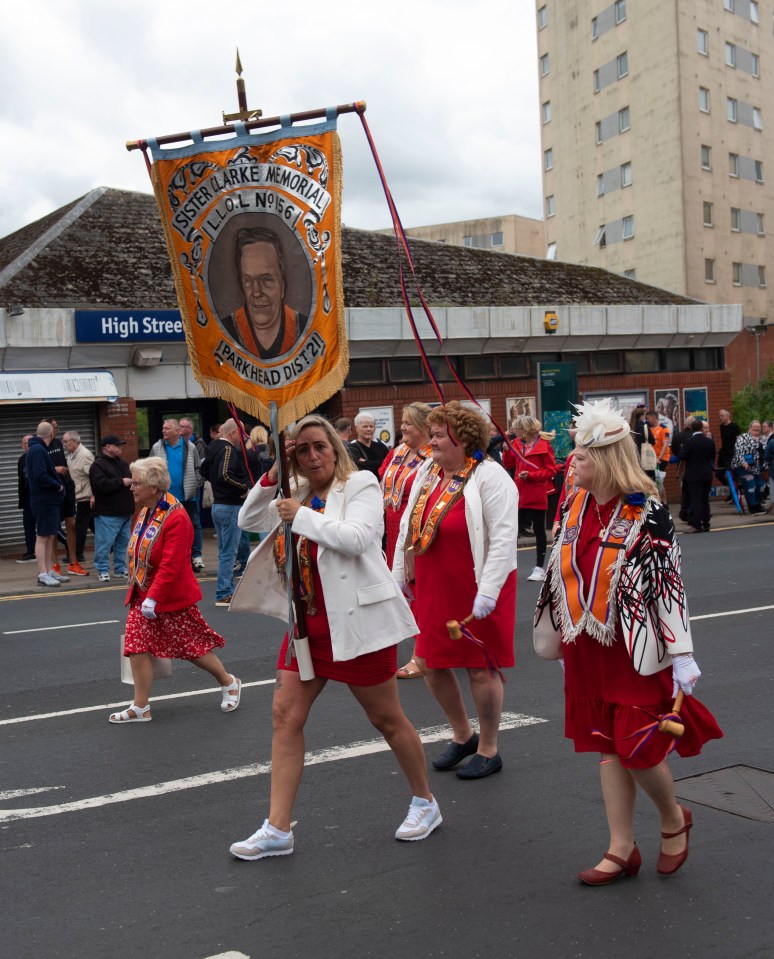 Women in red dresses and sashes carrying a memorial banner in a parade.
