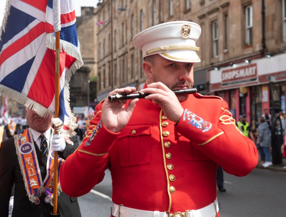 A man in a red uniform plays a flute during a parade.