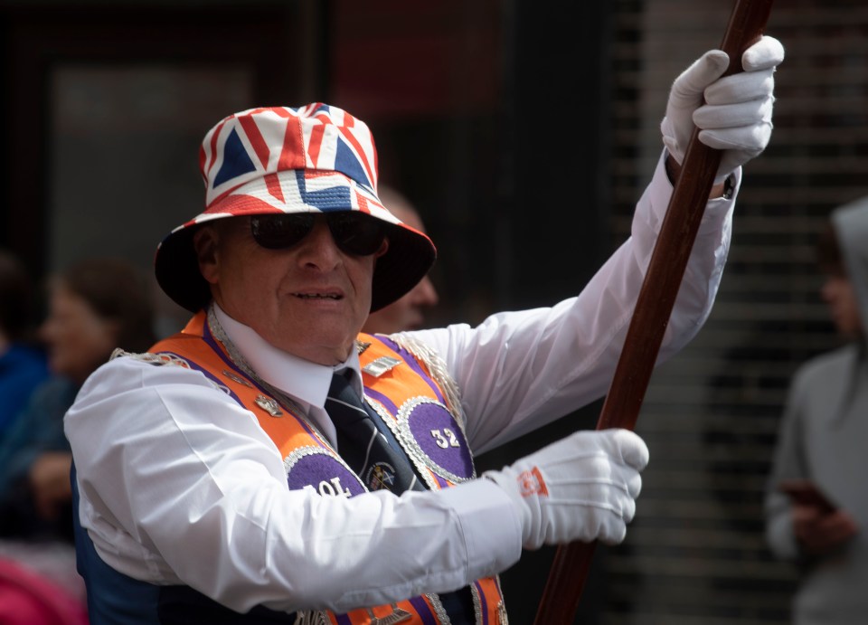 Man in Union Jack bucket hat and sash carries a staff.