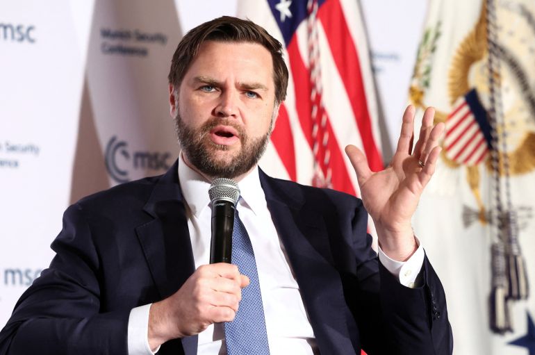 JD Vance lifts a hand in gesture and holds a microphone with the other as he speaks at a conference on stage.