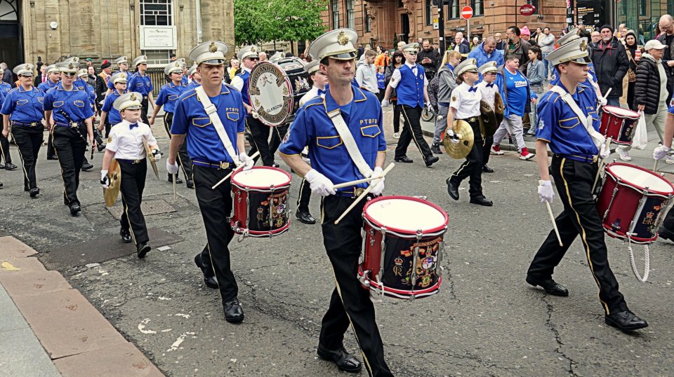 A marching band in blue uniforms and white hats parades down a city street.