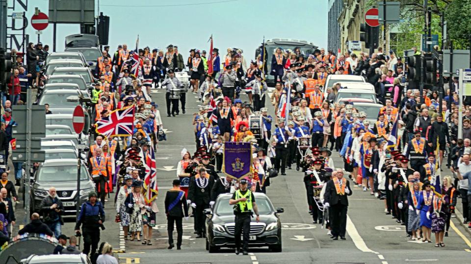 A large group of people in a parade, many wearing orange sashes, marching down a city street.