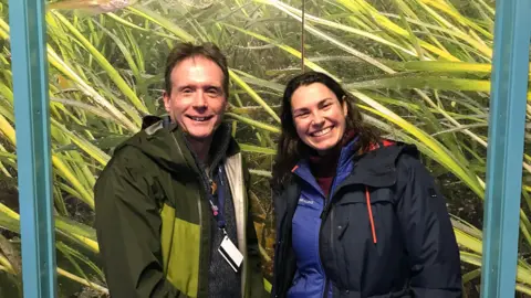 Martin Attrill wears a green coat, black shirt and lanyard and smiles at the camera, while Anna Turns wears a dark jacket and smiles widely. Behind them is an area of seagrass. 