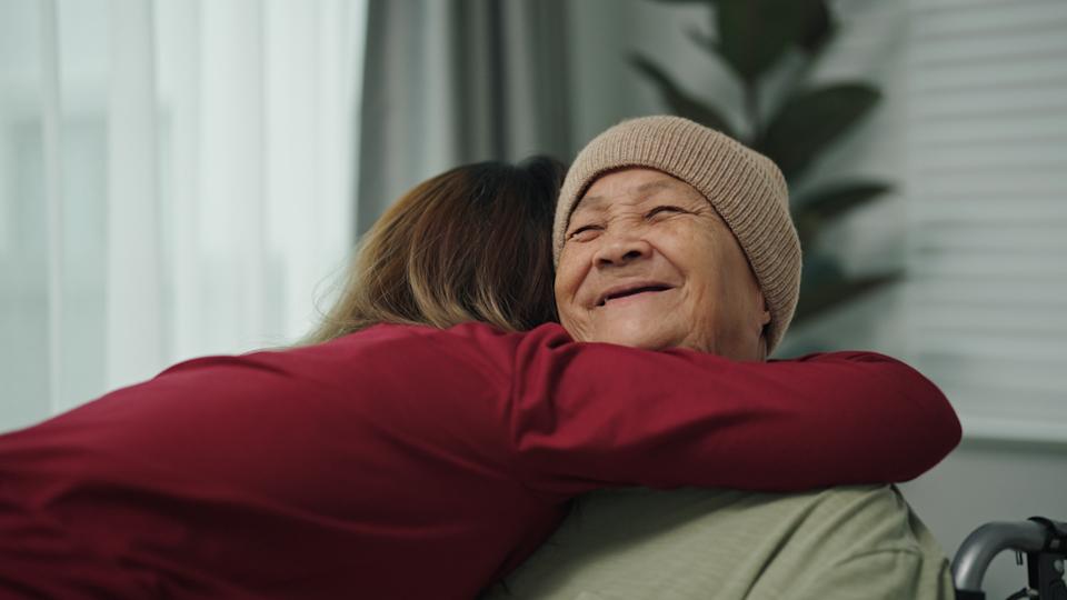 A person happily hugs an elderly person in a beanie, sitting in a wheelchair