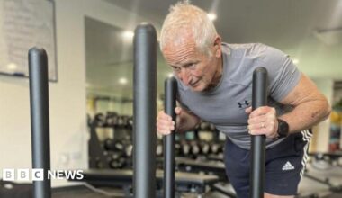 Peter Herbert in a grey tight-fitted shirt and blue shorts. His arms are grasping two vertical bars on an exercise sled which are almost touching his shoulders. His head is down and he is straining in his face and neck. He has white hair.