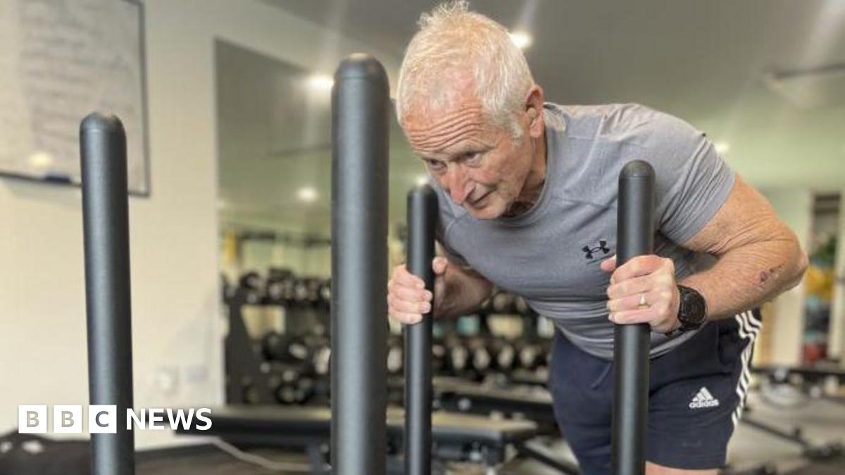 Peter Herbert in a grey tight-fitted shirt and blue shorts. His arms are grasping two vertical bars on an exercise sled which are almost touching his shoulders. His head is down and he is straining in his face and neck. He has white hair.