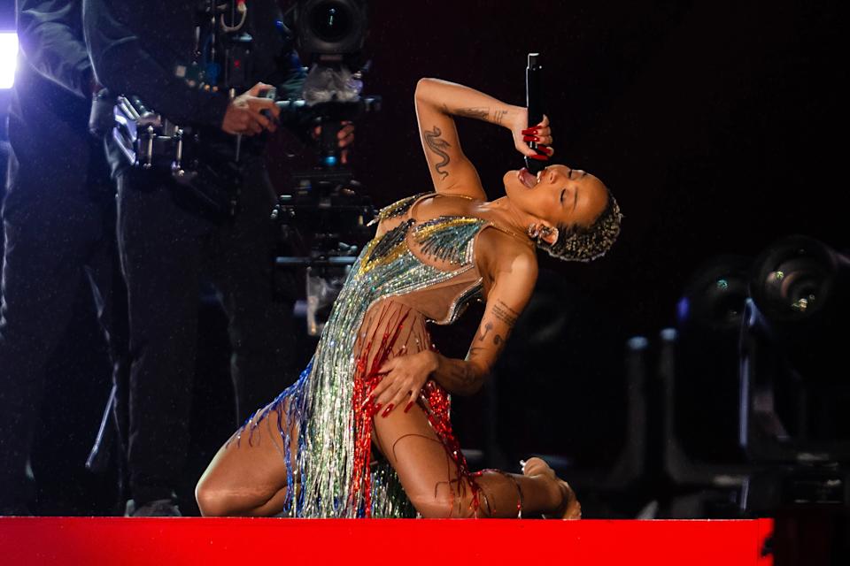A performer in a fringed, sequined dress poses dynamically on stage, holding a microphone, with camera operators in the background