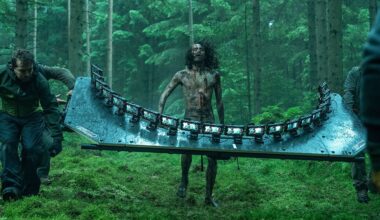 A person covered in mud stands in a forest while two crew members carry a large rig with multiple cameras mounted in a curved row; the scene appears to be from a film set.