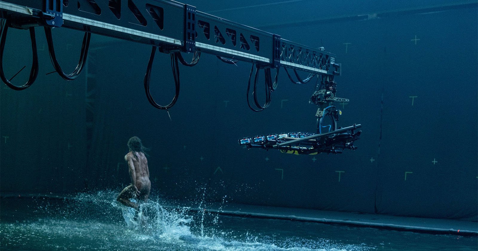 A person runs through shallow water on a film set, while a large camera rig on a suspended track films the scene. The background has blue screens with tracking markers for visual effects.