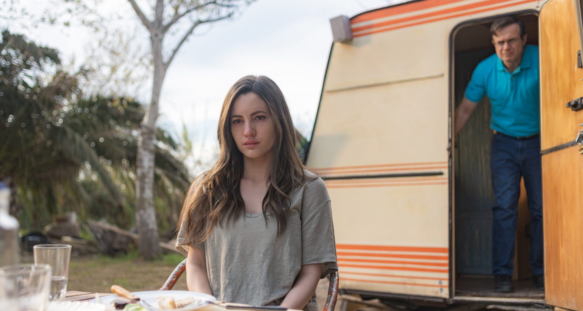 Maje (Ivana Baquero) sits in front of a caravan with Salva (Tristán Ulloa) in the doorway behind her in a scene from Netflix's &quot;A Widow's Game&quot;