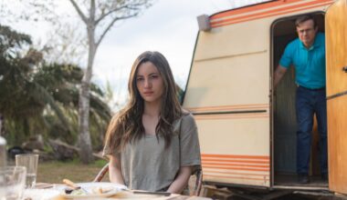 Maje (Ivana Baquero) sits in front of a caravan with Salva (Tristán Ulloa) in the doorway behind her in a scene from Netflix's &quot;A Widow's Game&quot;