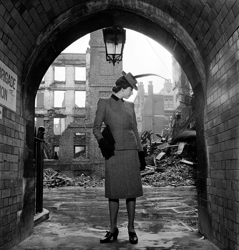 A woman in a tailored suit and hat stands under a brick archway, looking to the side, with the ruins of bombed buildings and rubble visible in the background.