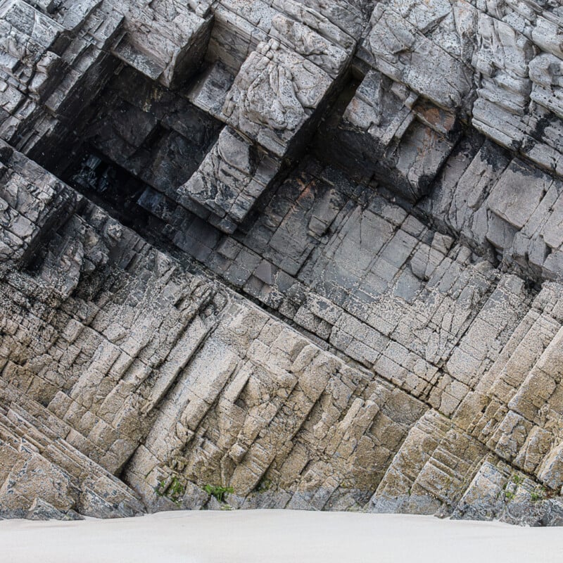 Jagged, layered rock formations with geometric patterns rise above a strip of pale sand, showcasing contrasting textures and angles in the natural stone.