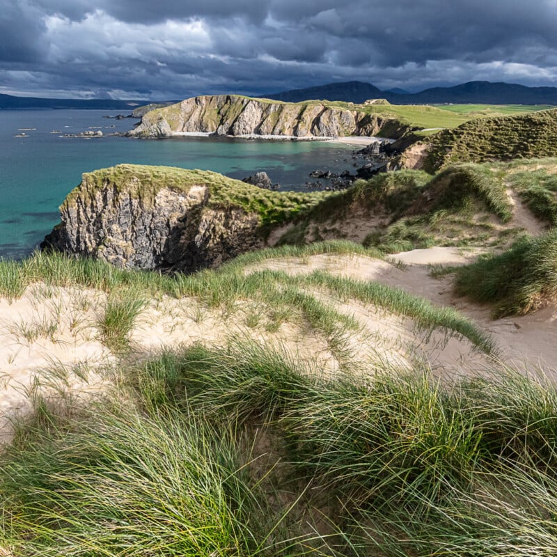 Grassy sand dunes overlook dramatic cliffs and turquoise sea under a cloudy sky at a rugged coastal landscape, with distant mountains visible on the horizon.