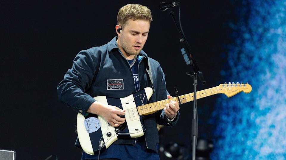 Sam Fender on stage, wearing a navy blue jacket and top. He is playing a black and white stripped guitar and stands in front of blue-black background.
