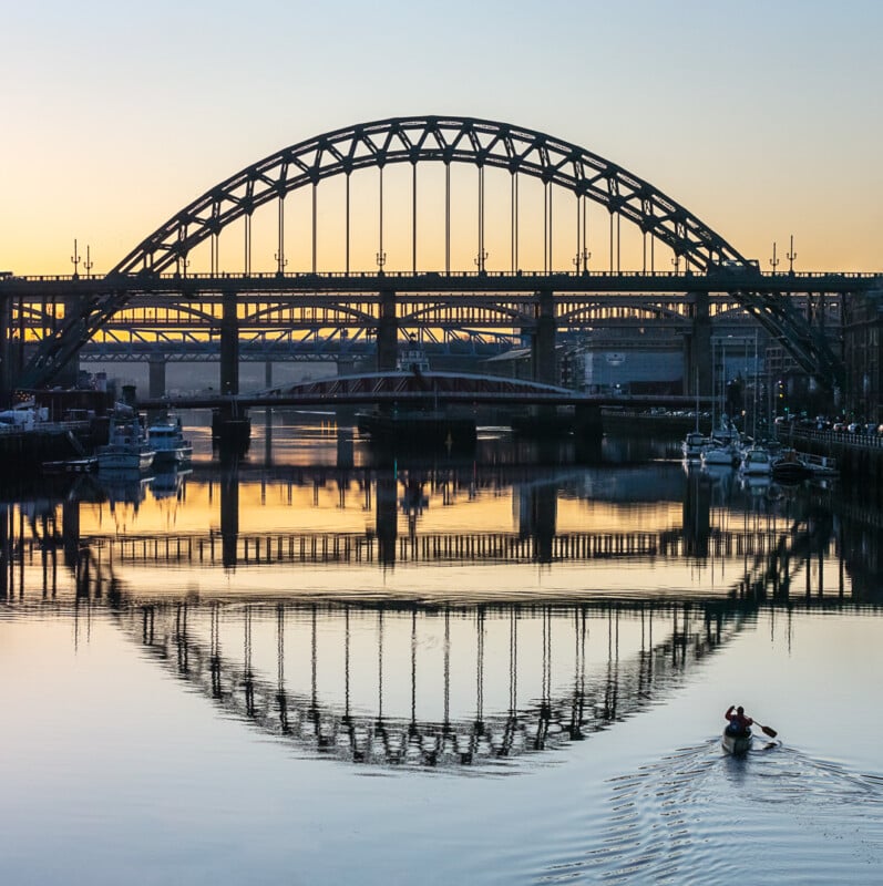 A person rows a boat on a calm river at sunset, with arched steel bridges reflected in the water and boats moored along the riverbanks.