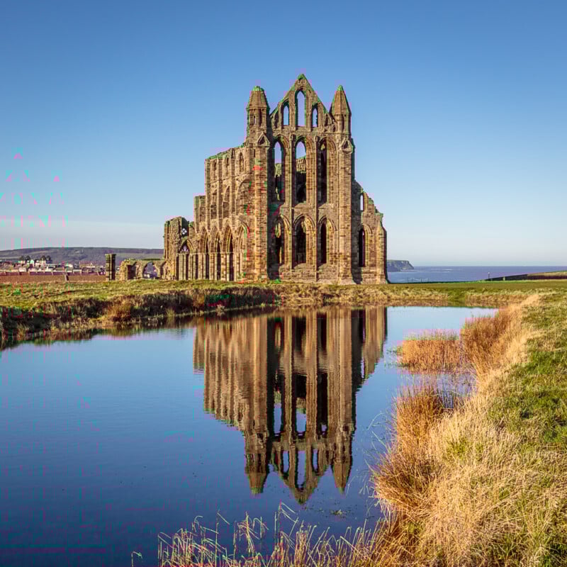 Ruins of a large stone abbey reflected in a still pond, with clear blue sky and grassy landscape surrounding the structure. Coastline and distant hills are visible in the background.