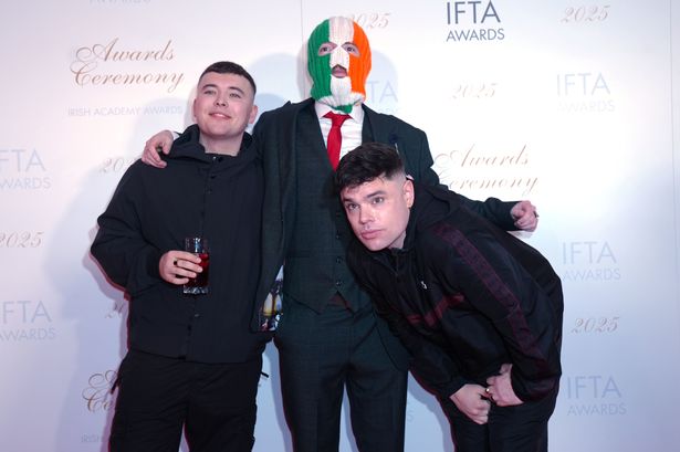 An image of three young men, two with dark hair wearing tracksuits, one wearing a suit and a balaclava in the colours of the Irish flag.