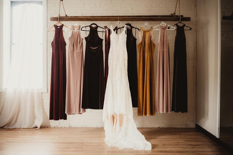 A row of elegant velvet gowns on hangers, with a white lace wedding dress centered, hanging from a wooden rack in a fashion display