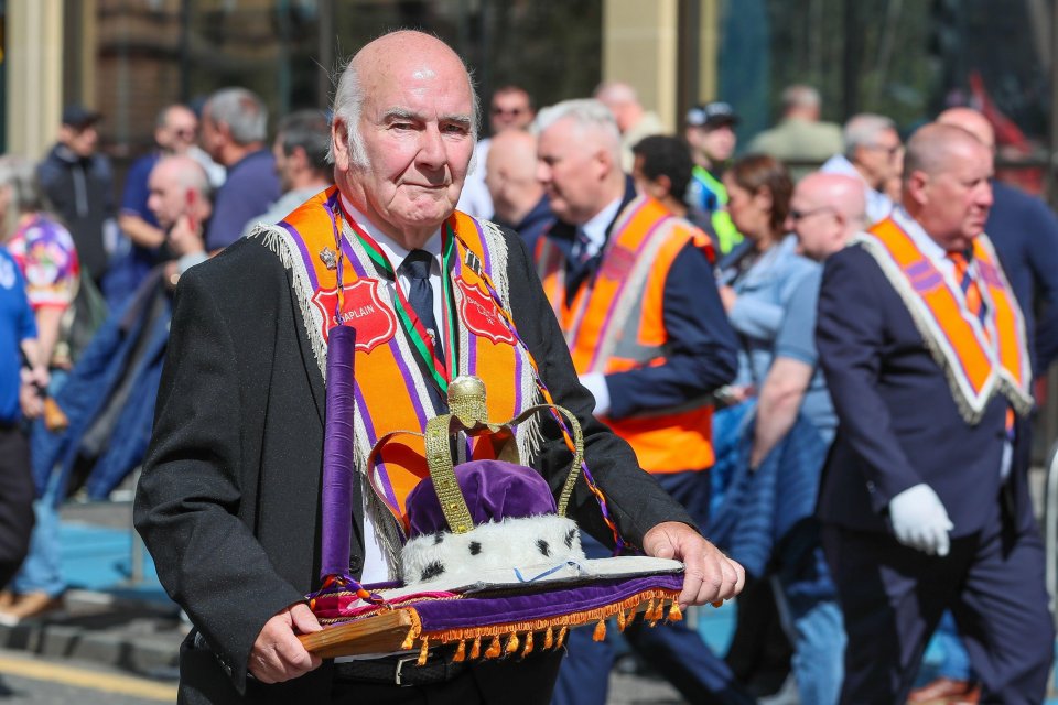 A man in an orange sash carries a crown and a book during a parade.