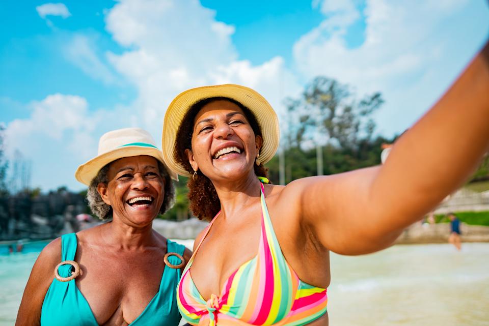Two people smiling and taking a selfie at a sunny beach, wearing hats and swimsuits. They appear joyful and relaxed