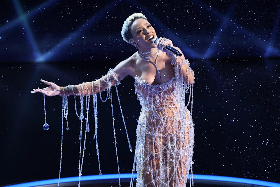 Singer on stage in intricate beaded gown, performing passionately with one arm extended, surrounded by a starry backdrop