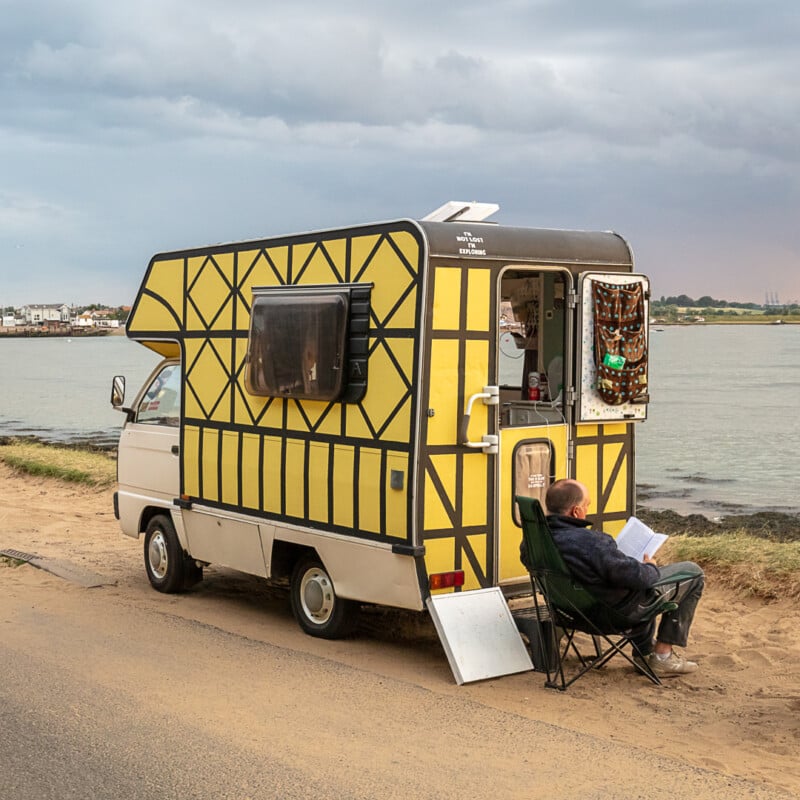 A person sits in a camping chair reading beside a small, yellow, half-timbered van parked by a river on a sandy roadside, with town buildings and cloudy skies in the background.