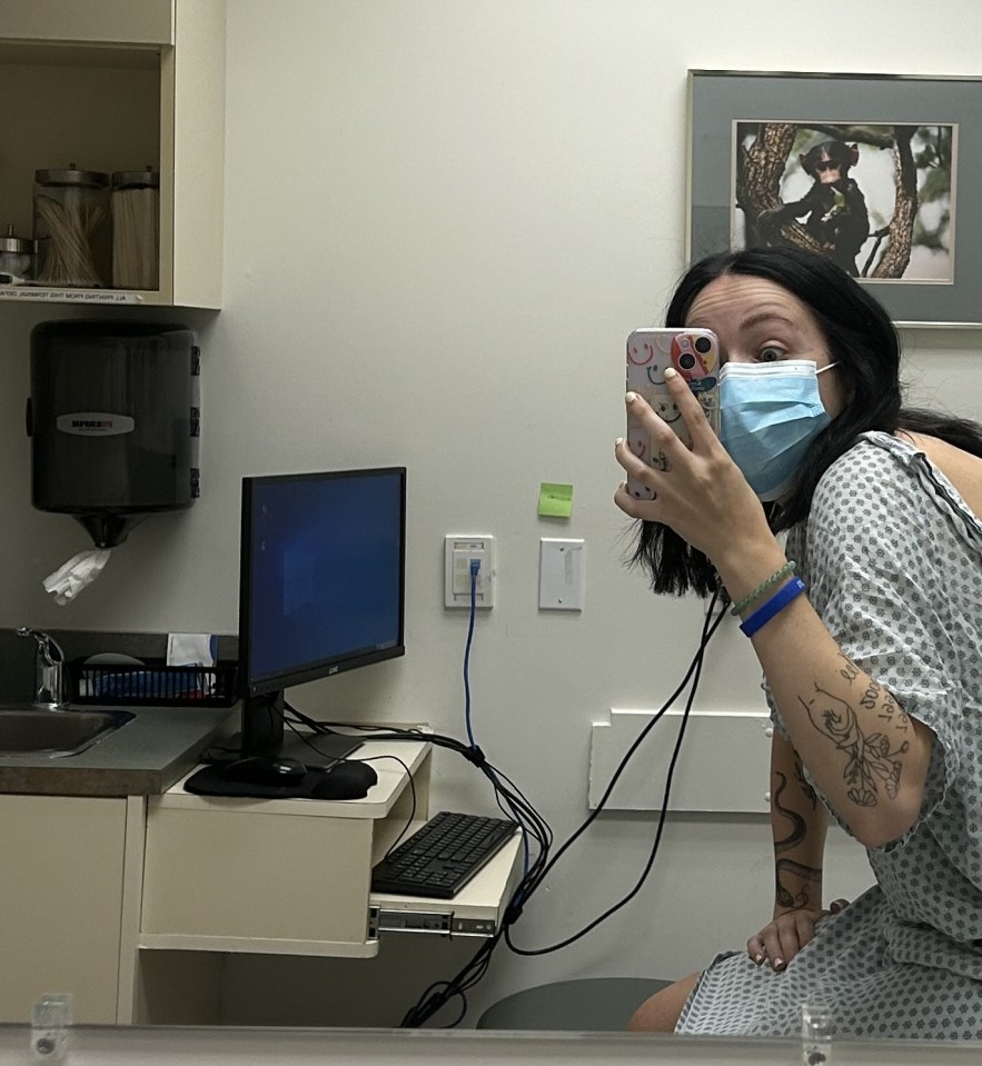 A young woman in a hospital gown, wearing a face mask, holds up her phone to take a selfie in a clinic waiting room.