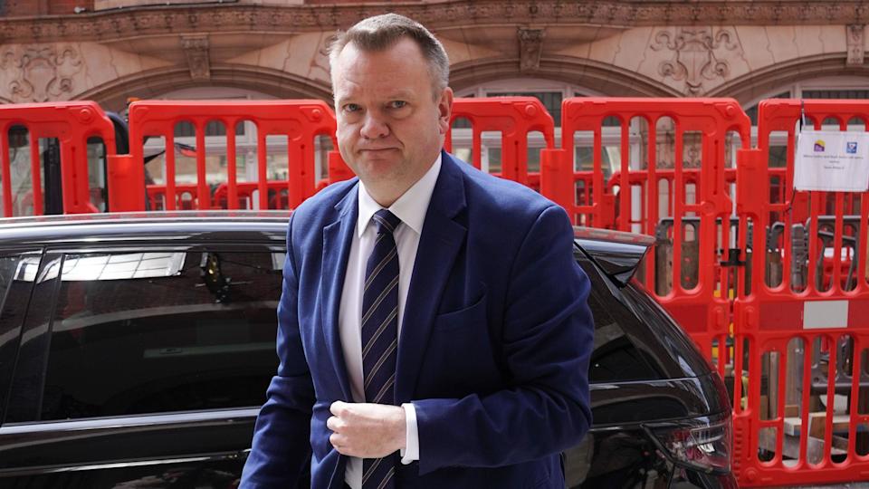 Nick Thomas-Symonds has short grey hair, he is wearing a navy suit and tie, with white shirt, and is pictured looking serious with a black car and red plastic railings  behind him