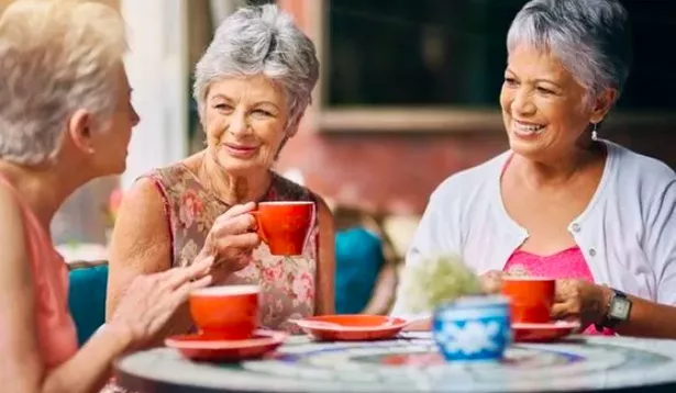 A trio of older women are sitting at a table smiling and drinking coffee 