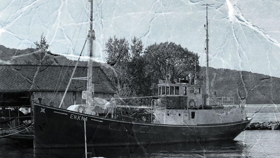 A black and white picture of the the M/K Erkna - a medium sized boat stationed by the shore with a building nearby. The picture appears to have been scanned so you can see lots of lines where it was folded. 