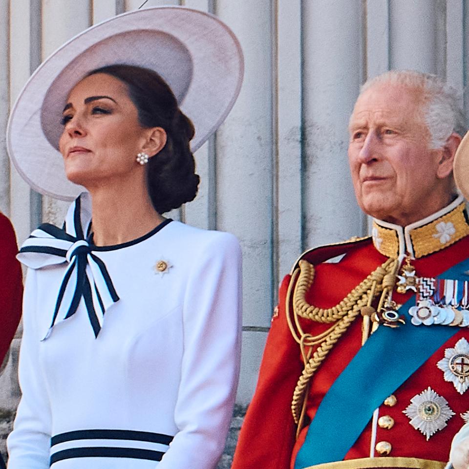  Prince William and King Charles wear red military uniform and Kate Middleton wears a white dress with navy accents and a matching hat on the Buckingham Palace balcony. 