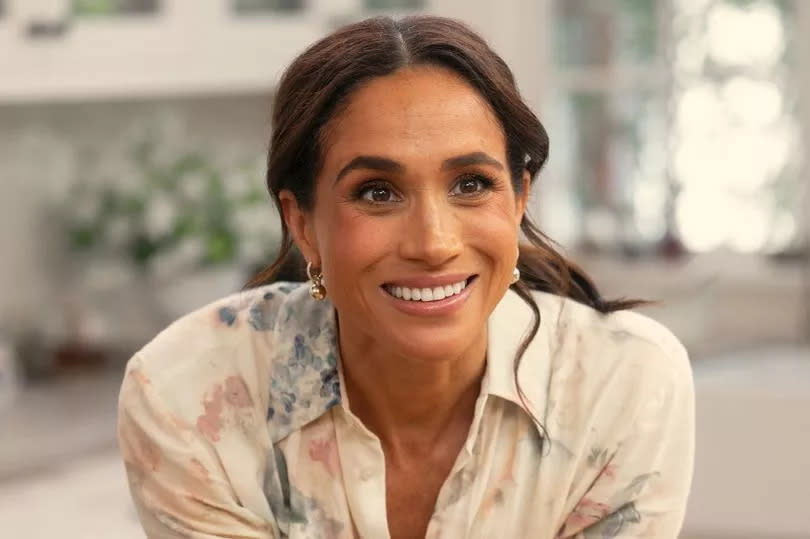 Meghan Markle in an off-white shirt leaning against a kitchen counter.