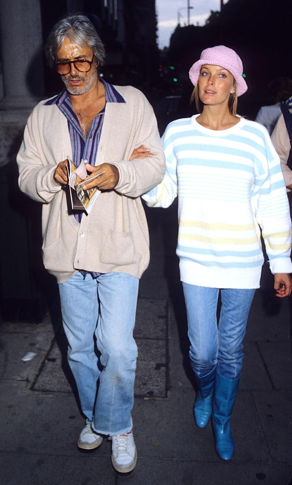 Two people walking. The man wears glasses, a cardigan, and jeans, holding a book. The woman wears a striped sweater, pink hat, jeans, and blue boots