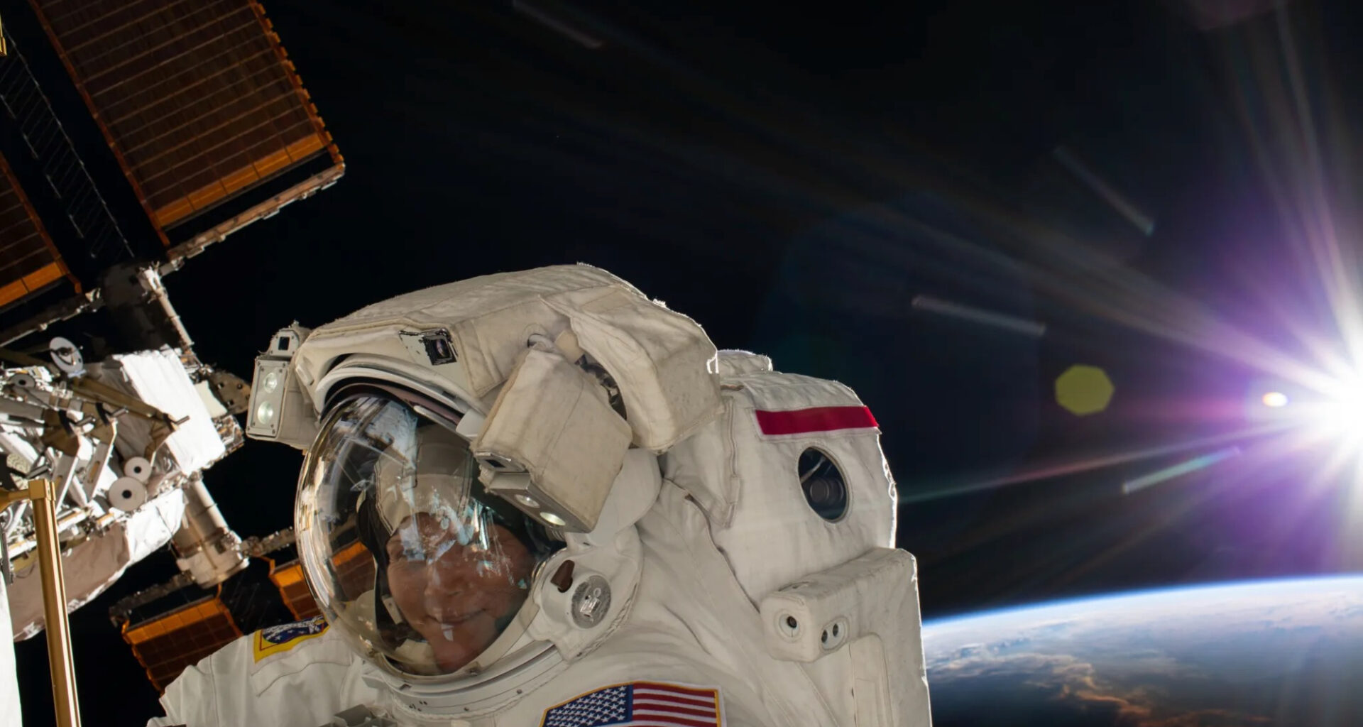 closeup of a smiling woman in a spacesuit conducting a spacewalk outside the international space station, with the sun and curve of earth in the background