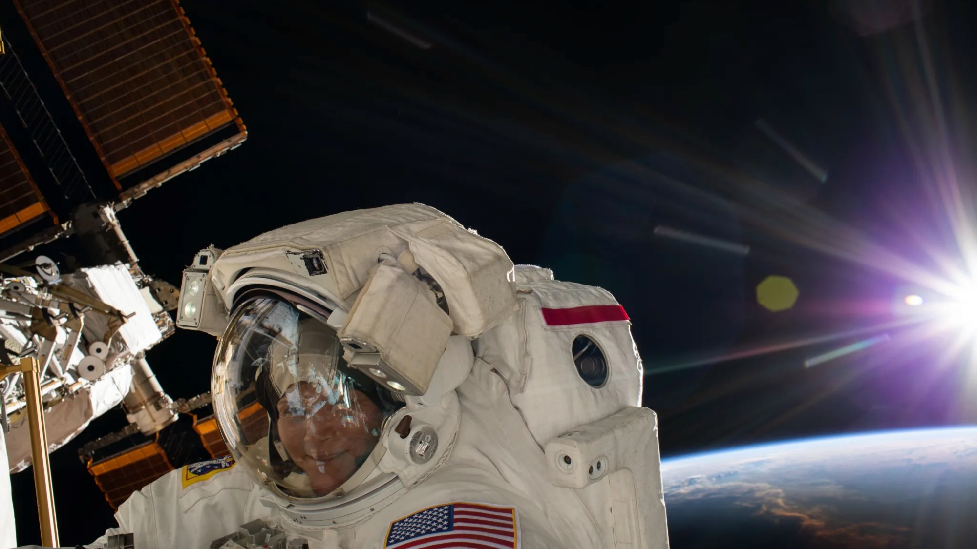 closeup of a smiling woman in a spacesuit conducting a spacewalk outside the international space station, with the sun and curve of earth in the background