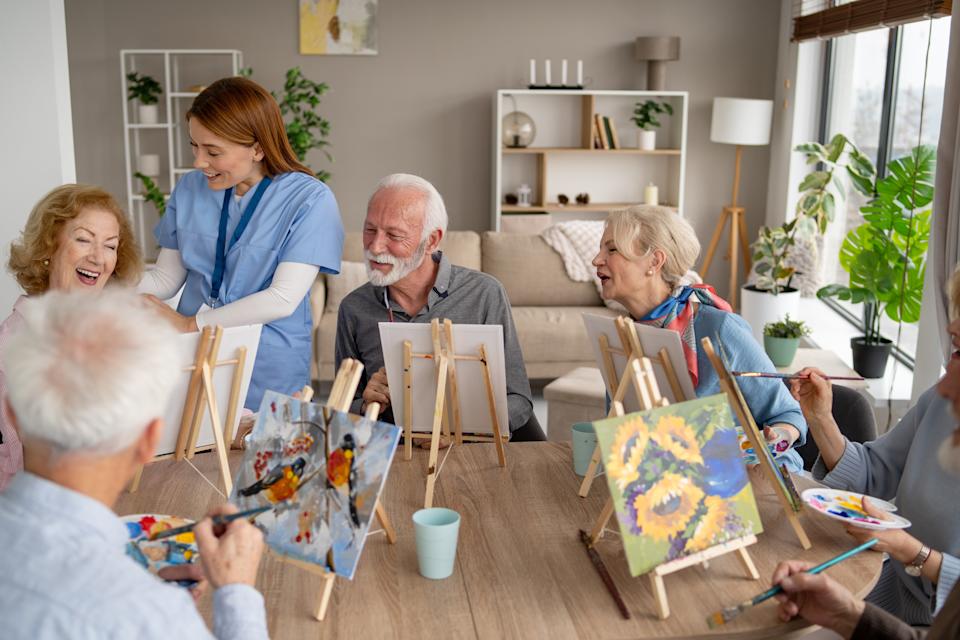 Group of older adults enjoying a painting class with an instructor in a cozy room. They are smiling and focused on their canvases