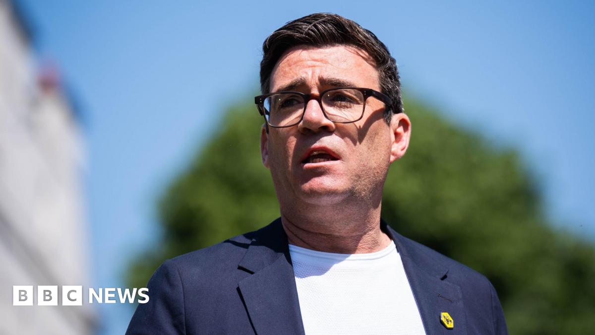 Andy Burnham has dark brown hair, dark brown glasses, a white T-shirt and blue jacket.