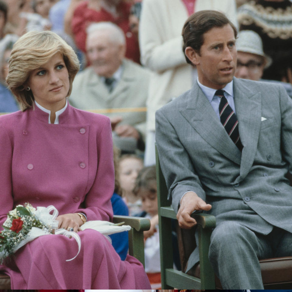  Prince Charles wearing a gray suit and Princess Diana in a pink suit holding flowers sitting in chairs next to each other with a crowd behind them. 