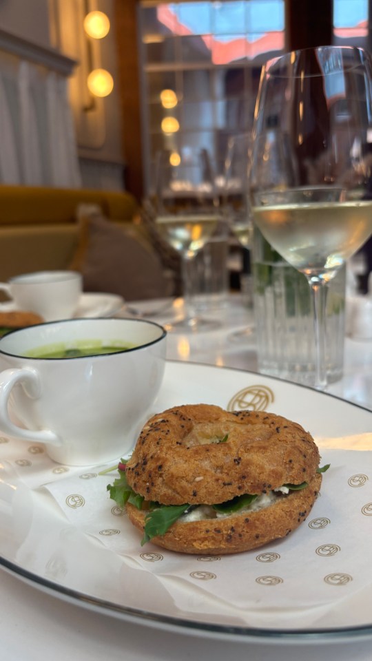 Bagel sandwich with cream cheese and greens on a plate with soup and wine in the background.
