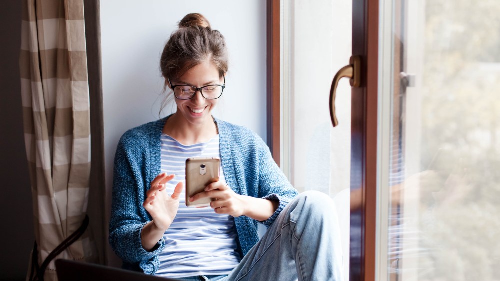 Holiday shopping online. Young woman makes purchases in the Internet. Freelancer pays by using smartphone on Black Friday. Happy girl is enjoying buying gifts and sitting by window with laptop at home