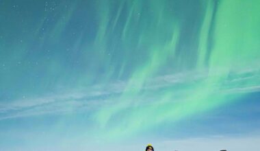 A group of 13 people standing together on snow-covered ground, dressed in warm winter clothing, with the aurora glowing in the sky above them. Some are kneeling, while others stand in front of them, all facing the camera and smiling.