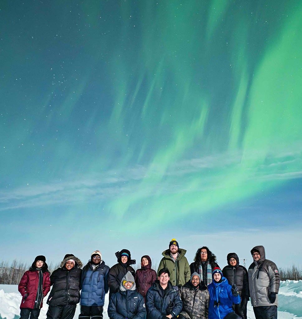 A group of 13 people standing together on snow-covered ground, dressed in warm winter clothing, with the aurora glowing in the sky above them. Some are kneeling, while others stand in front of them, all facing the camera and smiling.