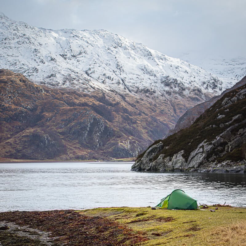 A green tent is pitched on grassy ground by a calm lake, with rugged hills and snow-capped mountains in the background under a cloudy sky.