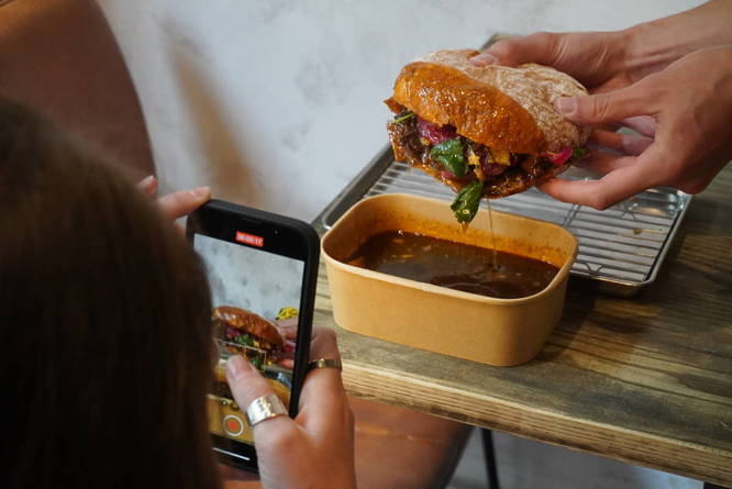Someone taking a photo of a sandwich being dipped into a pot of sauce
