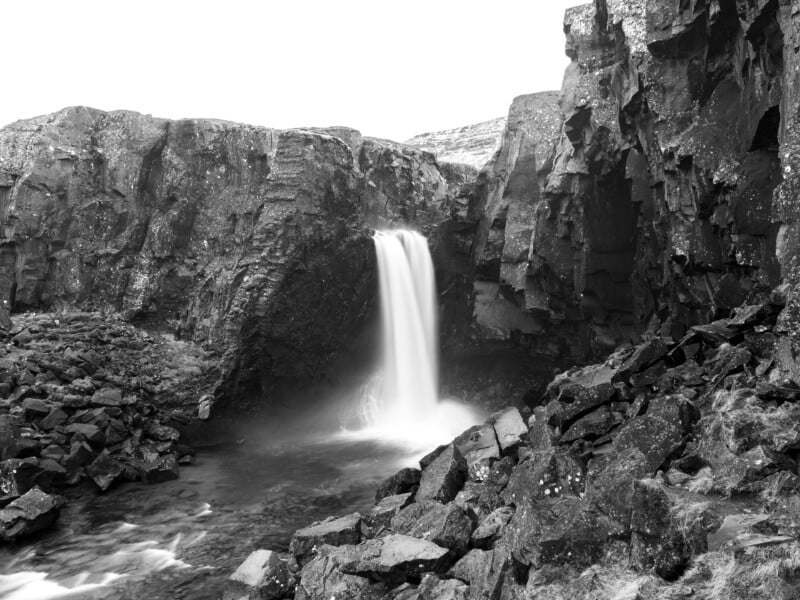 Black and white photo of a waterfall cascading between rugged, rocky cliffs into a pool below, surrounded by jagged rocks and boulders. The water appears silky due to a long exposure.