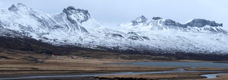 Snow-capped jagged mountains rise above a brown grassy plain with a small cluster of houses near a winding river under a cloudy, overcast sky.