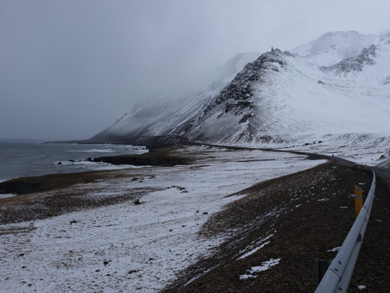 A winding coastal road runs alongside a snow-dusted mountain slope, with the ocean to the left and low clouds shrouding the peaks, creating a cold, misty atmosphere.
