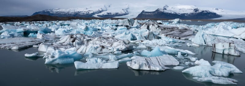 Large chunks of ice float in a glacial lagoon, with snow-capped mountains and a glacier in the background under a cloudy sky.